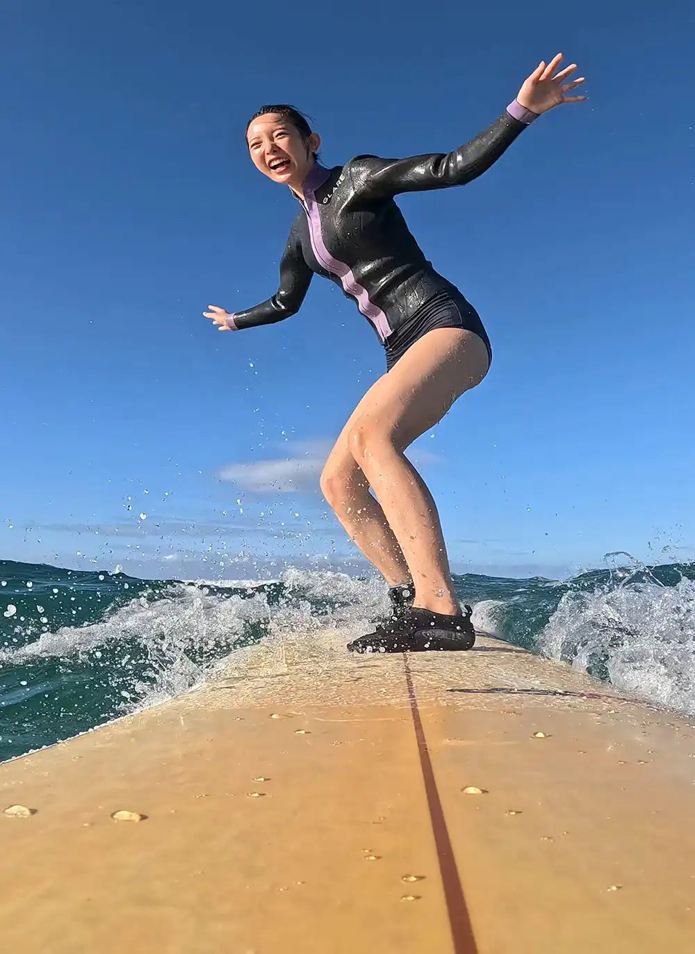 ワイキキの海で笑顔でサーフボードに立ち、波に乗るスクール参加者。CLIPSハワイのサーフレッスン中に撮影