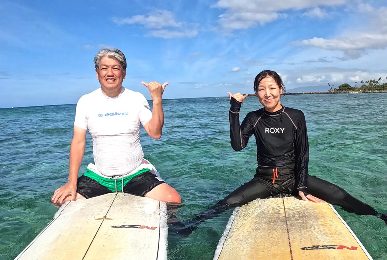 ハワイの海でサーフボードに座り、シャカサインで笑顔を見せる二人の受講者。CLIPSハワイのサーフレッスン中に撮影された写真
