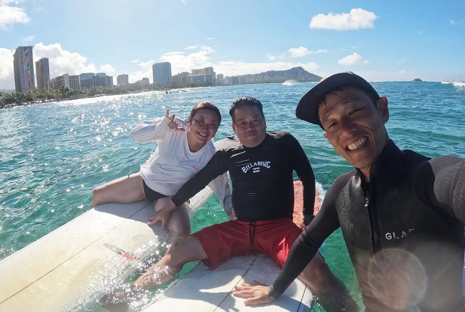 ハワイの海でサーフボードに座り、シャカサインをしながら楽しむレッスン参加者と日本人インストラクター。CLIPSハワイの少人数制レッスン写真。