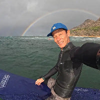 ハワイの海でサーフボードに座り、背後に虹がかかる中で笑顔を見せる日本人インストラクターの紹介写真。