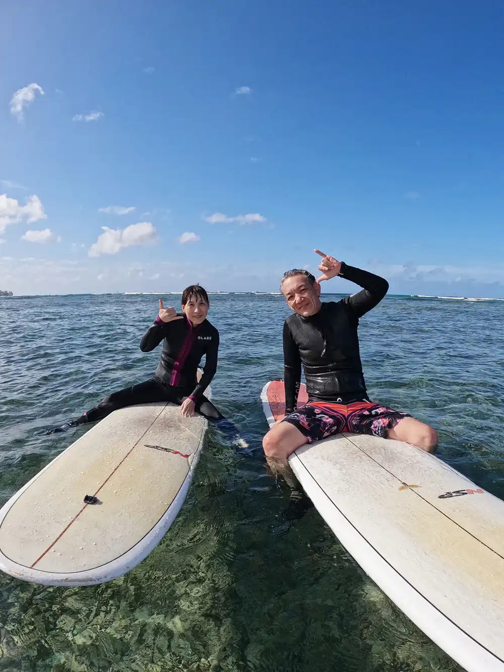 ハワイの浅瀬でサーフボードに座り、シャカサインをして楽しむカップルの受講者。CLIPSハワイの初心者向けサーフレッスンで撮影された写真。
