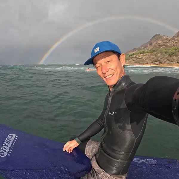 ハワイの海でサーフボードに座り、背後に虹がかかる中で笑顔を見せる日本人インストラクターの紹介写真。